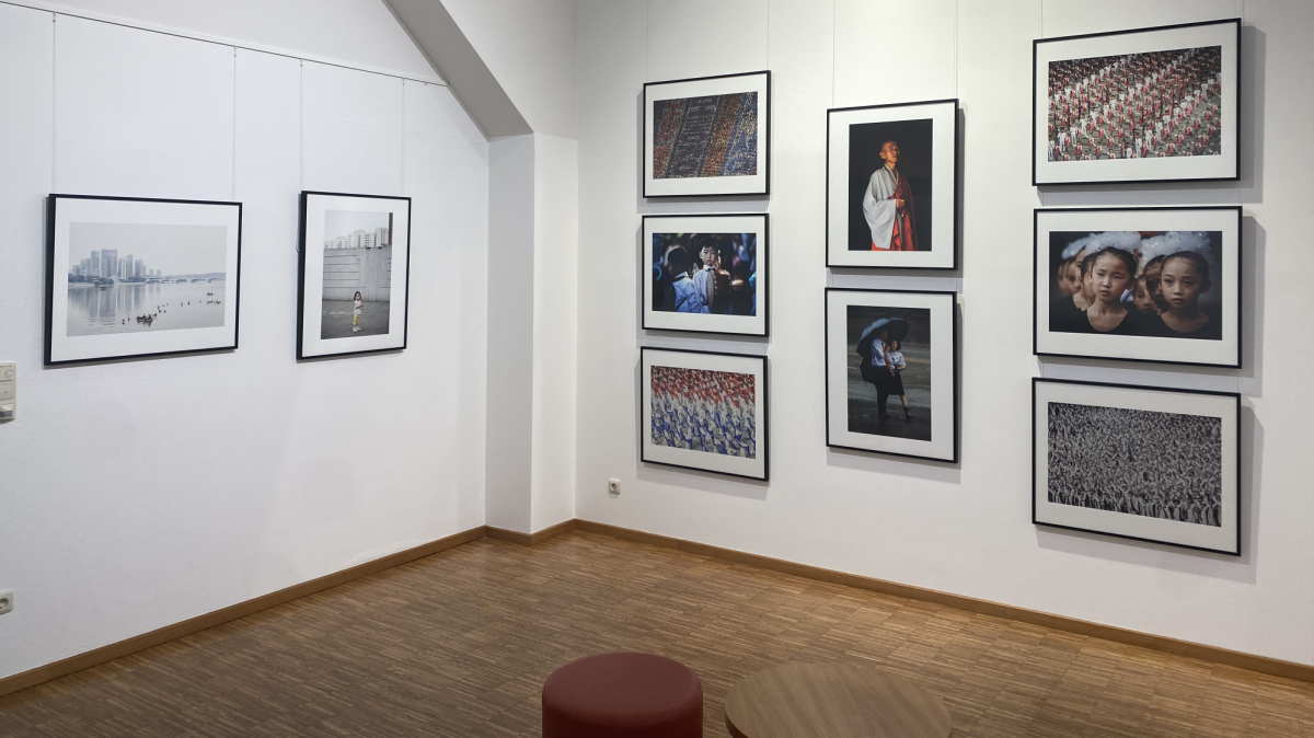 In einem Raum mit Parkettboden hängen großformatige Fotos in schwarzen Rahmen an den weißen Wänden. Auch ein Tischchen mit Hocker stehen im Raum.