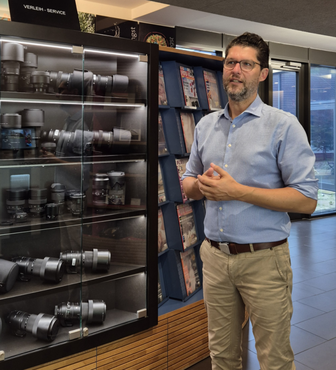 Ein Mann mit Brille und Bart, in hellblauem Hemd und beiger Hose steht vor einem Schaukasten voller Fotokameras und Objektiven und erklärt etwas.
