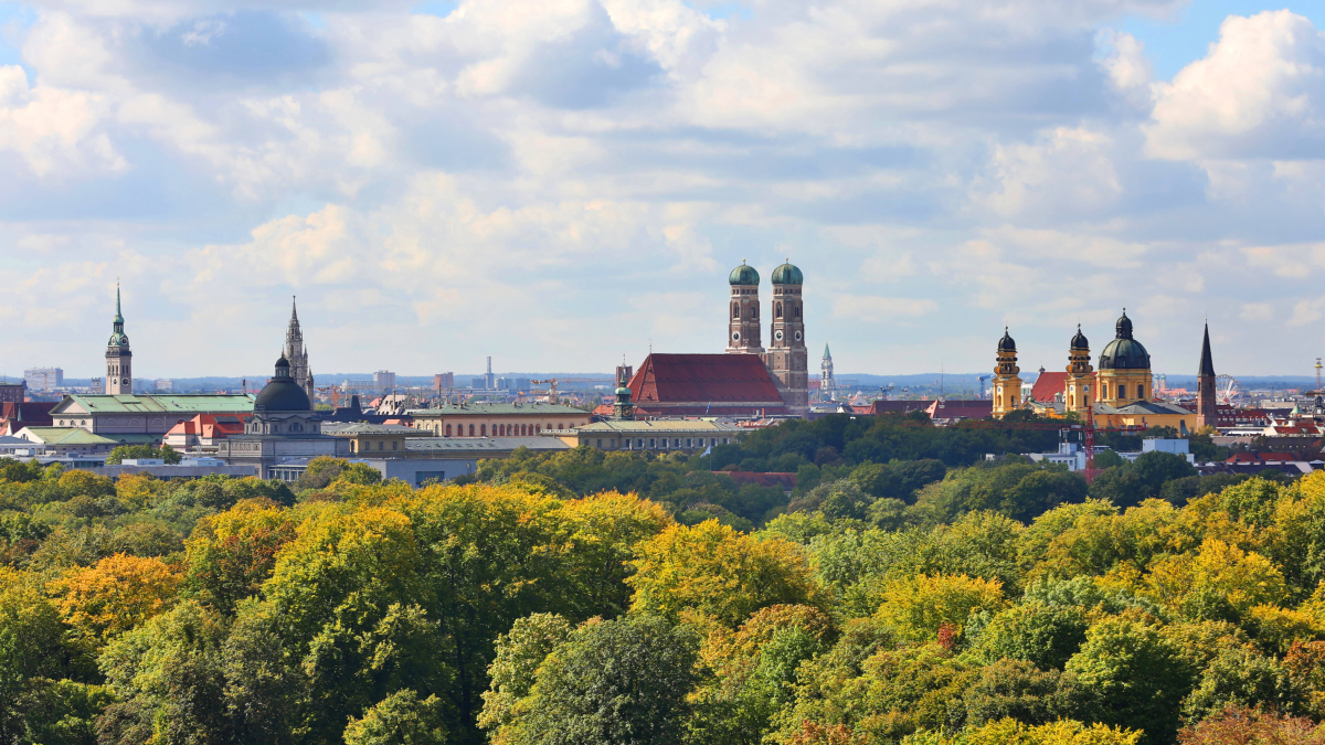 Häuserdächer und Kirchtürme zwischen Baumkronen und sonnig bewölktem Himmel.
