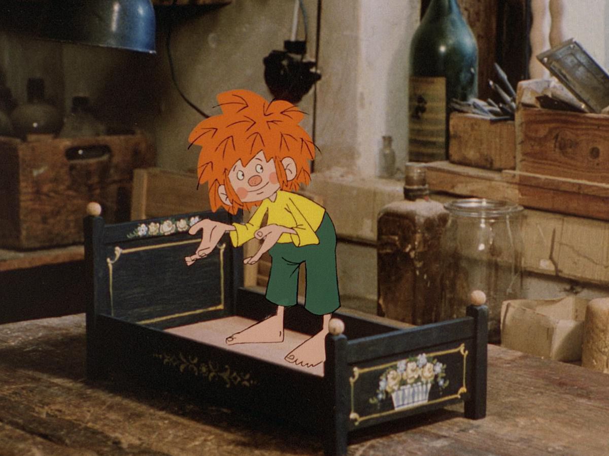 Der kleine Kobold Pumuckl in seiner grünen Hose, gelbem Shirt und knallroten Wuschelhaaren steht in einem kunstvoll bemalten Puppenbett aus Holz. Im Hintergrund sieht man Werkzeuge des Schreiners. Der Pumuckl ist eine gezeichnete Figur in einer real gefil