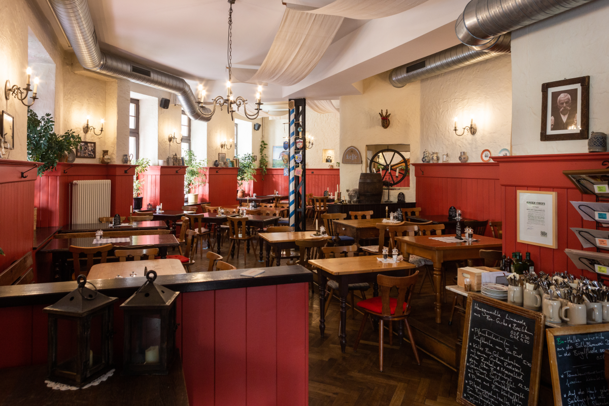 Ein großer Gastraum mit rot lackierten, holzgetäfelten Wandverkleidungen. Die Tische und Stühle sind aus massivem Holz mit gedrechselten Beinen. An den Wänden sind Lampen im Stil von Kerzenhaltern, einige Jagdtrophäen und alte Fotos in Rahmen angebracht. 