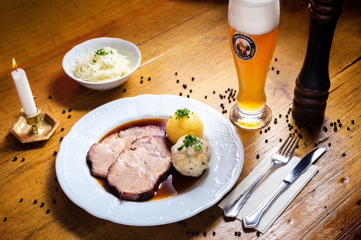 Auf einem weißen Teller sind zwei Scheiben Schweinsbraten in Saft und mit zweierlei Knödel (Kartoffel- und Semmelknödel) angerichtet, mit Schnittlauch und Pfefferkörnern garniert. daneben Besteck auf Serviette, eine Schale mit Kraut, ein Glas Bier, eine h