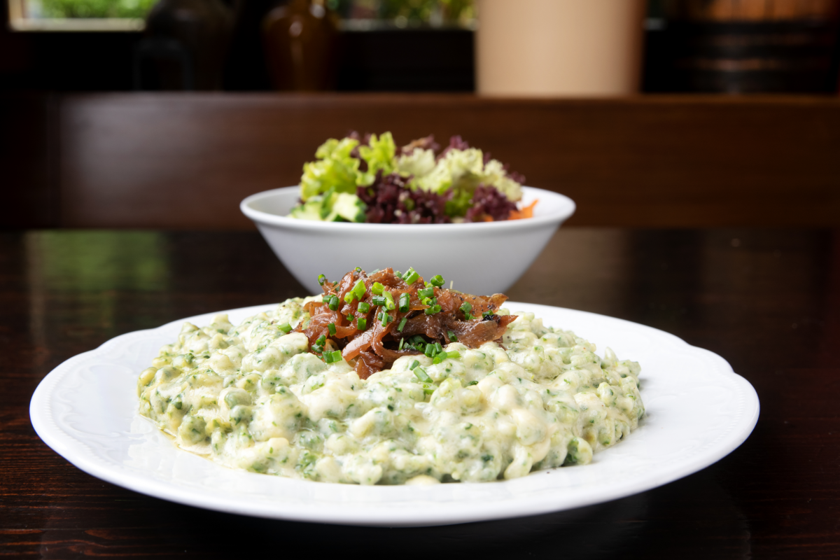 Spinatspätzle in Käsesauce auf einem Teller angerichtet, mit gerösteten karamellisierten Zwiebelringen und Schnittlauch garniert. Daneben eine Schale mit gemischtem Salat.