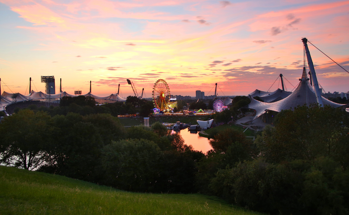 Blick vom Olympiahügel auf den Park mit Stadion und Vergnügungspark mit beleuchtetem Riesenrad. Das Zeltdach überspannt das Areal in weitem Bogen, dazwischen dicht belaubte Bäume. Der leicht bewölkte Abendhimmel dahinter ist vom Sonnenuntergang in pastell