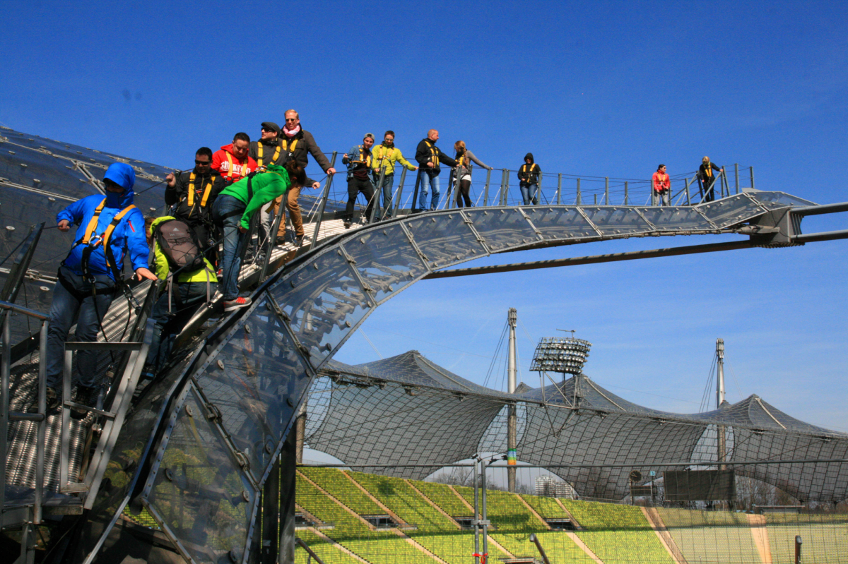 Eine Gruppe von Menschen wandert über einen Steg mit Geländer über die stählerne Zeltdachkonstruktion des Olympiastadions. Sie tragen dabei Gurte und sind gesichert. Der Himmel im Hintergrund ist tiefblau.