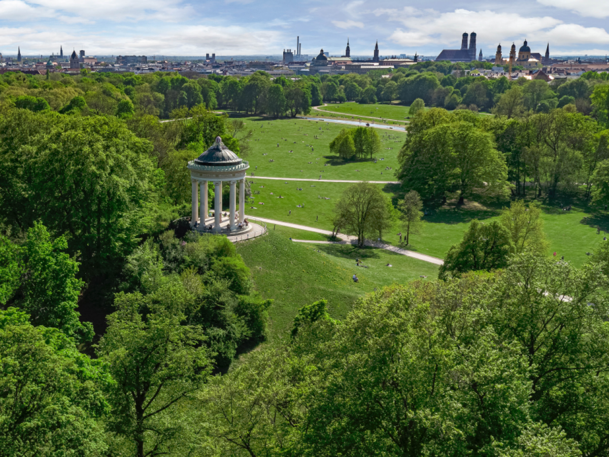 Blick auf einen weitläufigen Park: Viel dicht belaubter Baumbestand, sich schlängelnde Wege, auf einem Hügel ein Monopteros (antiker Rundtempel aus einer kreisförmigen Säulenreihe mit Dach ohne Wände) und am Horizont die Skyline der Münchner Innenstadt.