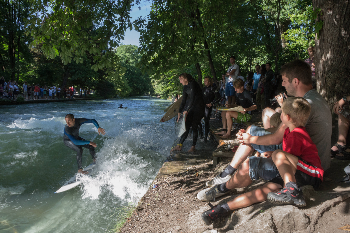 Auf einem Bach, der viel türkisblaues, weiß schäumendes Wasser führt, surft ein Mann im Neoprenanzug auf einem weißen Surfbrett. Weitere Surfer warten am gemauerten Ufer, auf den Felsen sitzen Zuschauer verschiedenen Alters.