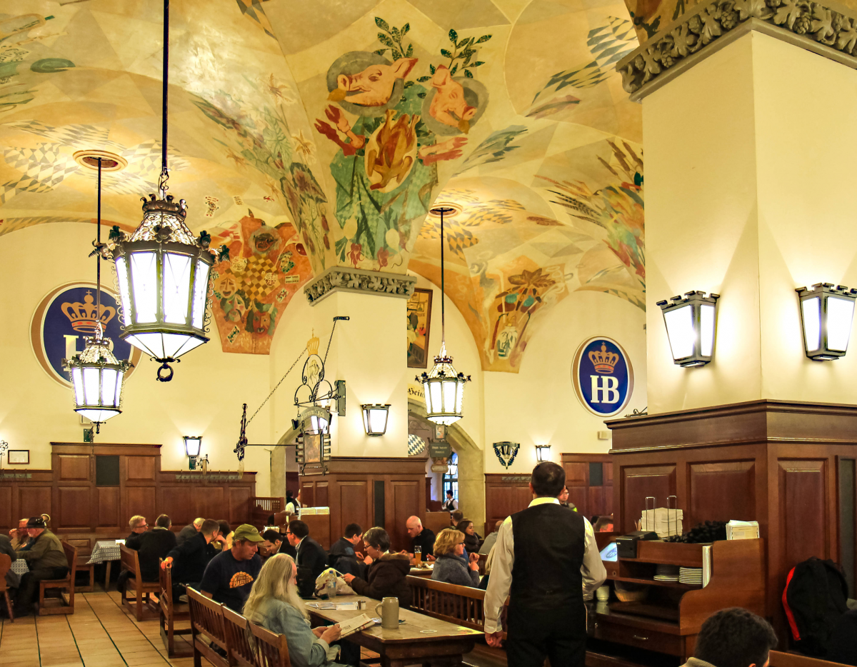 Der riesige Gastraum, überspannt von einem kunstvoll bemalten Deckengewölbe auf mit Stuck verzierten Säulen. Rustikale Laternen an den Wänden und von der Decke hängend. Mit dunklem Holz vertäfelte Wände, Gäste sitzen auf Holzbänken an langen Tischen.
