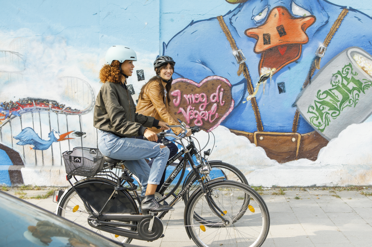 Zwei Frauen auf schwarzen Fahrrädern, an der Wand hinter ihnen zeigt ein großflächiges Mural eine scheinbar betrunkene, blaue Comic-Ente in Trachtenlederhose, mit einem Maß Bier und einem Lebkuchenherz mit der Zucker-Aufschrift: 