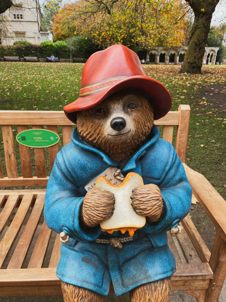 Eine Paddington-Figur auf einer Parkbank in London: Der Bär trägt einen roten Schlapphut, einen blauen Mantel und hält ein Marmeladesandwich zwischen den Pfoten. Im Hintergrund sieht man eine herbstliche Parkwiese, Bäume und ein Haus.