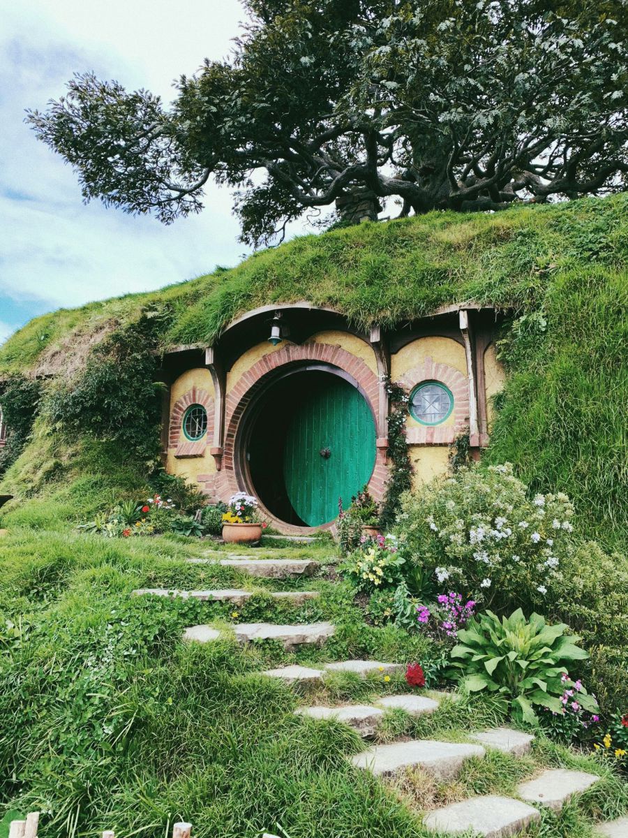 Hobbit-Höhle: In einem grasbewachsenen Erdhügel ist ein Eingangsbereich mit rundem Tor und kleinen, runden Fenstern eingelassen. Ein paar Steinstufen führen hinauf, daneben ein kleiner, gepflegter Vorgarten. Oben am Hügel wurzelt ein mächtiger, knorriger 