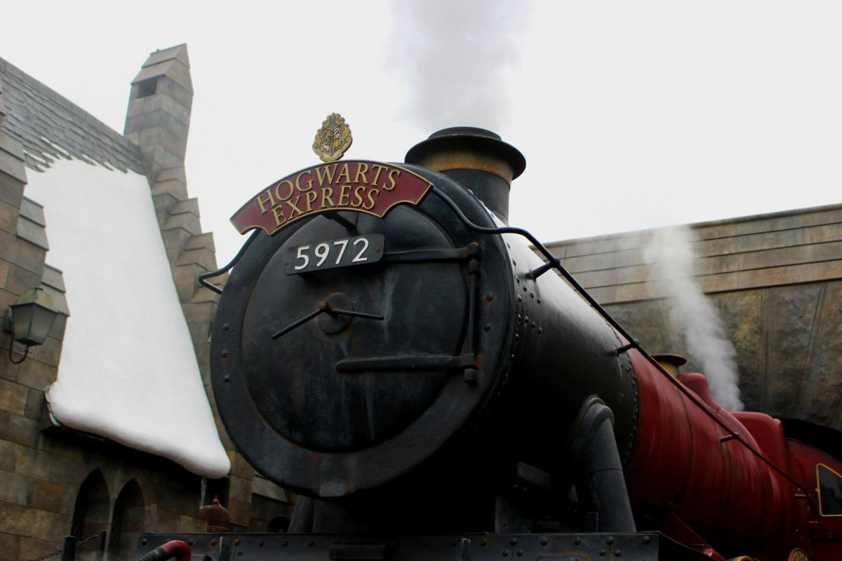 Die schwarz-rote Dampflok des Hogwarts Express in Frontansicht. Auf einem bordeauxrotem Schild mit goldenem Hogwarts-Wappen steht in goldener Schrift 