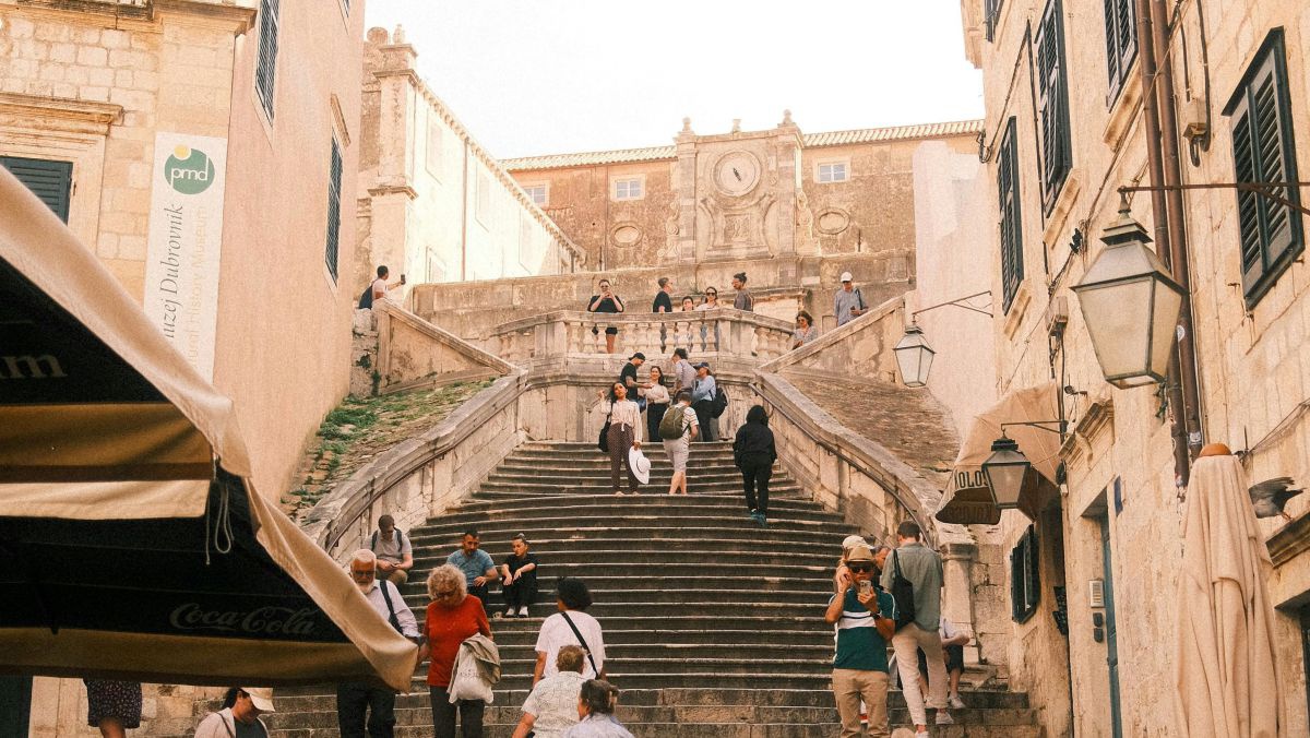 Zwischen hohen Stadhäusern mit dunkelgrünen Fensterläden führt eine breite, lange, teilweise schlängelnde, steinerne Treppe eine Anhöhe hinauf. Ganz oben steht ein dominantes Gemäuer mit großer Uhr über dem Eingang.