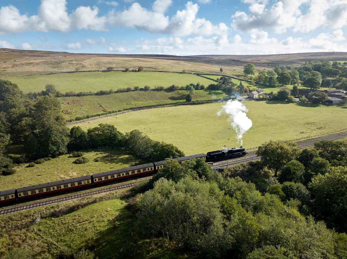 Eisenbahn mit Dampflok fährt auf einer geraden Strecke durch grüne, hügelige Landschaft. Himmel ist blau mit weißen Schäfchenwolken.