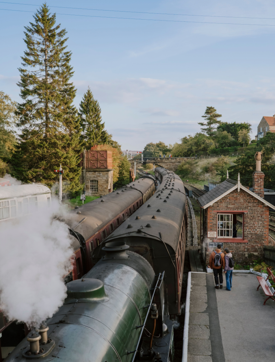 Kleiner Bahnhof mit Backstein-Wartehäuschen, auf den Gleisen beider Richtungen fahren soeben alte Eisenbahnzüge mit Dampfloks ein. In der Umgebung grüne Landschaft mit vereinzelt Backsteinhäuschen und hohen Bäumen.