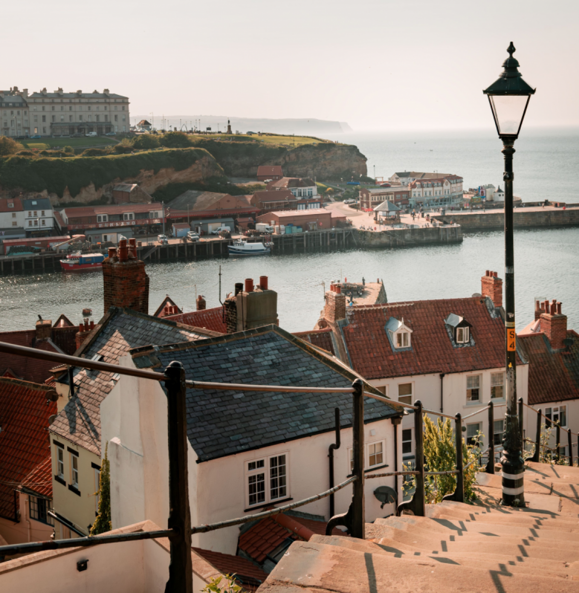 Blick von einer sonnenbeschienenen Steintreppe mit schmiedeisernem Geländer und Laterne hinunter auf die Hafenbucht. Die Häuser sind weiß gestrichen und haben graue und rote Dächer.