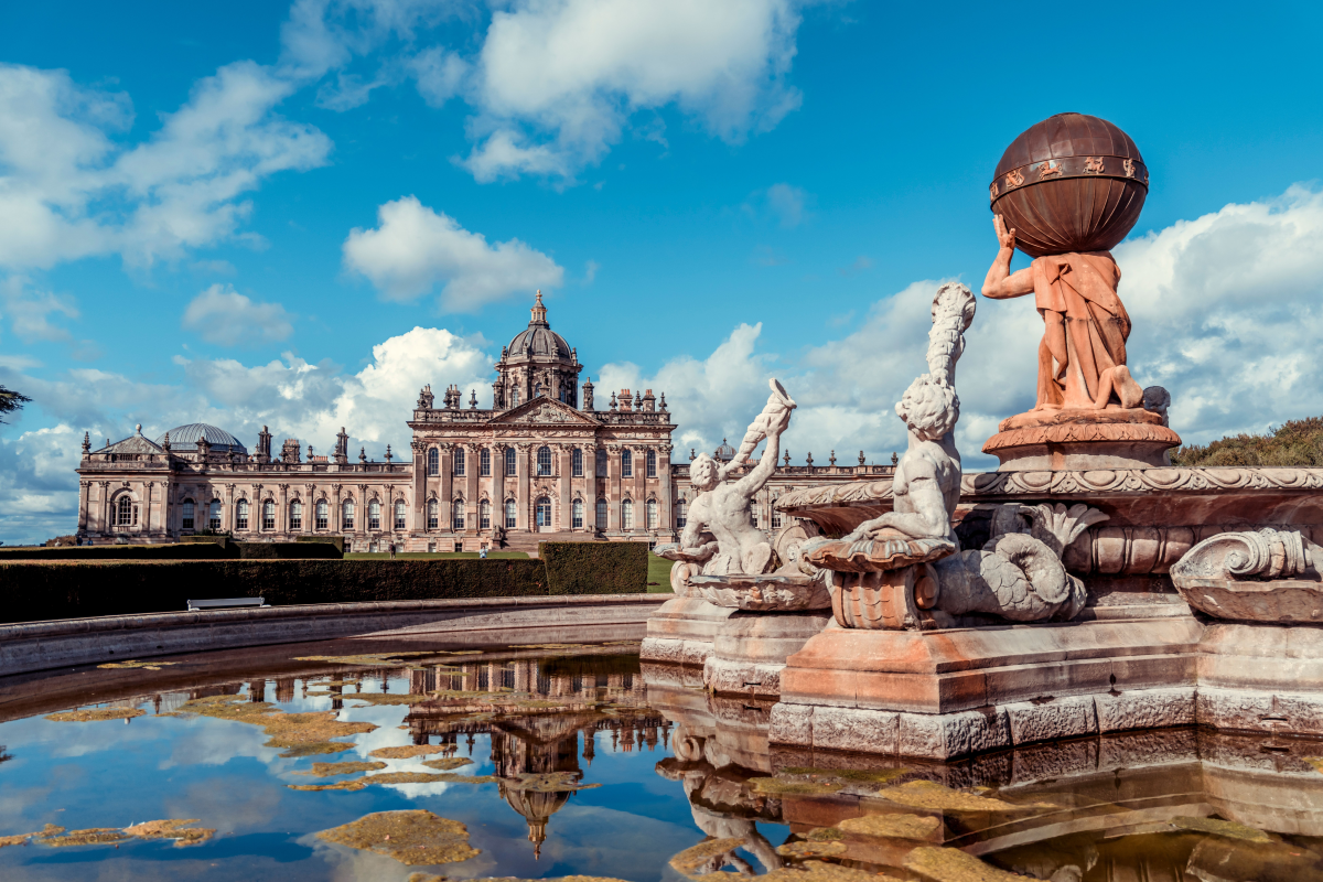 Im Vordergrund ein großer barocker Springbrunnen mit Marmorstatuen. Im Wasser spiegelt sich der blaue, mit weißen Wölkchen bevölkerte Himmel und das dahinterliegende, riesige Prunkschloss.