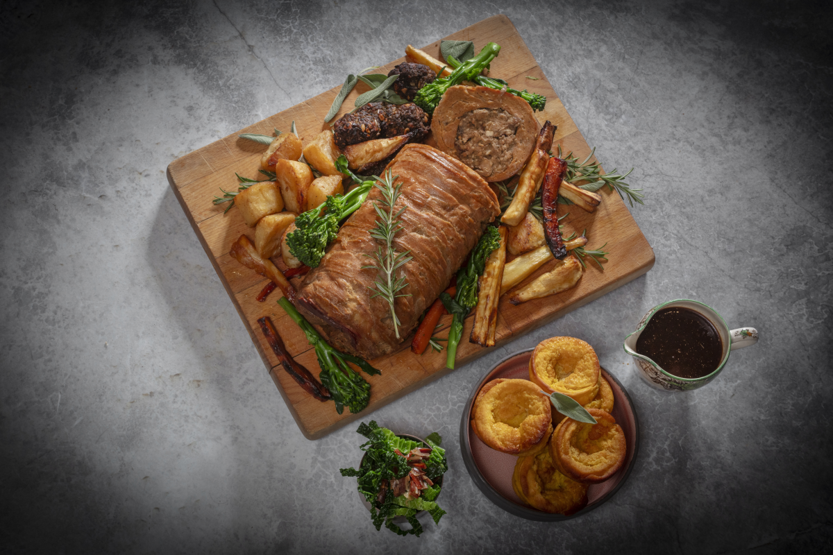 Veganer Sonntagsbraten angerichtet mit Bratkartoffeln, Gemüse und Kräutern auf einem Holzbrett.
