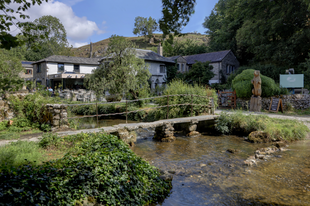 Kleine steinerne Brücke mit Eisengeländer über einem seichten Flüsschen, die Ufer sind Efeubewachsen. Zwischen Bäumen und Büschen eine Anordnung mehrerer kleiner Gebäude, Steinmauern, ein Gastgarten. Dahinter grüne Hügel.