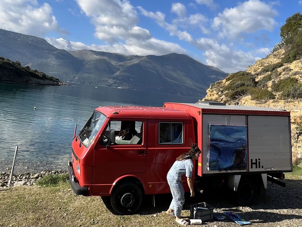 Am steinigen Ufer in einer Bucht parkt ein altes Feuerwehrauto, eine Frau im Overall bückt sich um Werkzeug aus einer Kiste. An der Ladewand des Fahrzeugs hängt eine bemalte, quadratische Leinwand.