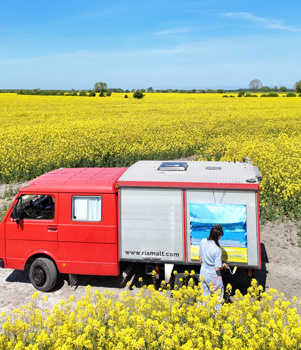 Zwischen grün-gelben Rapsfeldern und knallblauem Himmel parkt ein rotes Feuerwehrauto. Am Ladekontainer hängt eine Leinwand, auf der eine Frau gerade ein Bild der Landschaft malt. 