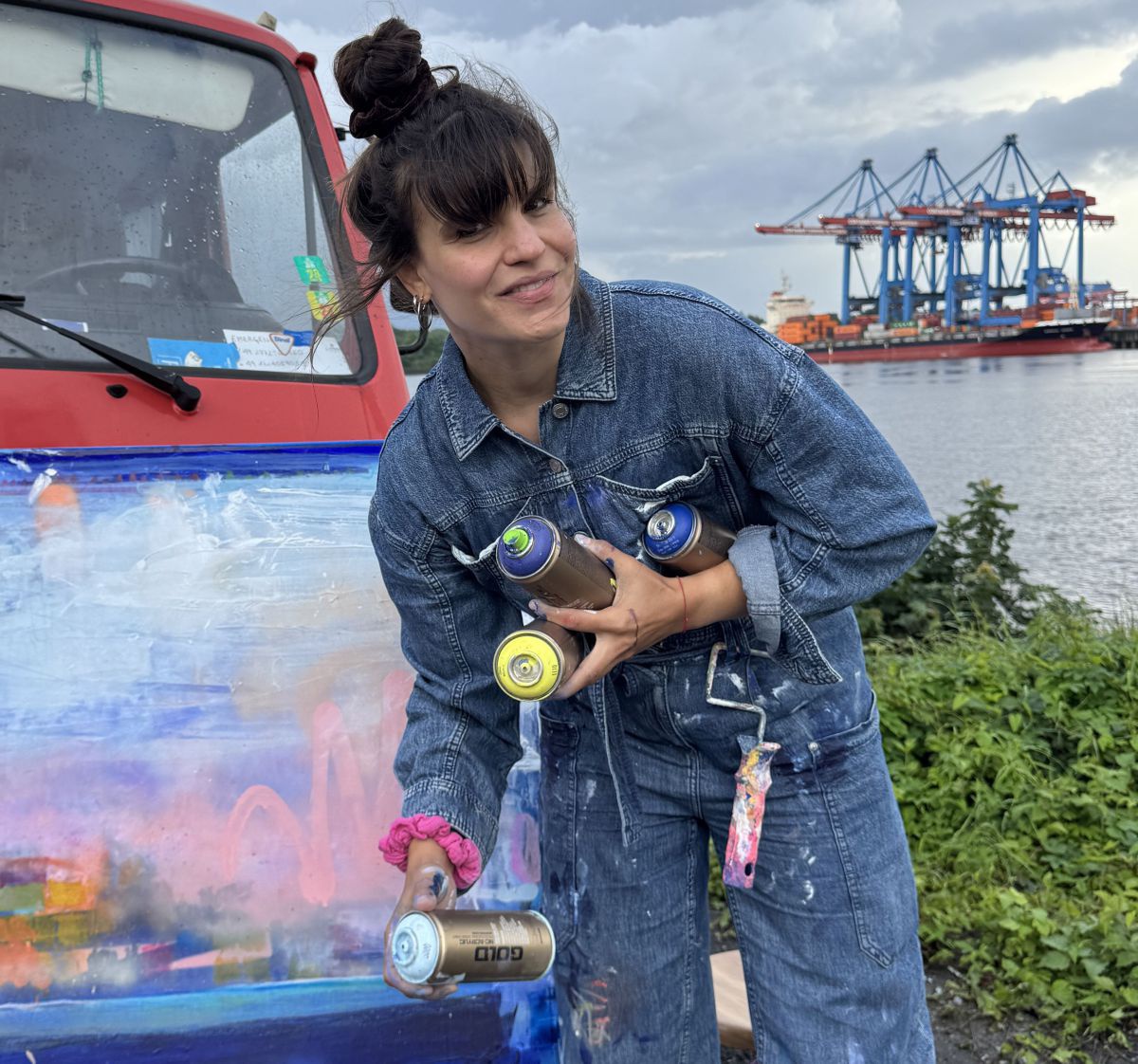 Eine junge Frau im Jeansoverall hält mehrere Farbspraydosen und Walzen in Händen, neben ihr lehnt eine bereits bemalte Leinwand an der Motorhaube eines roten LKW. Im Hintergrund ein Hafen mit Industriegerüsten.