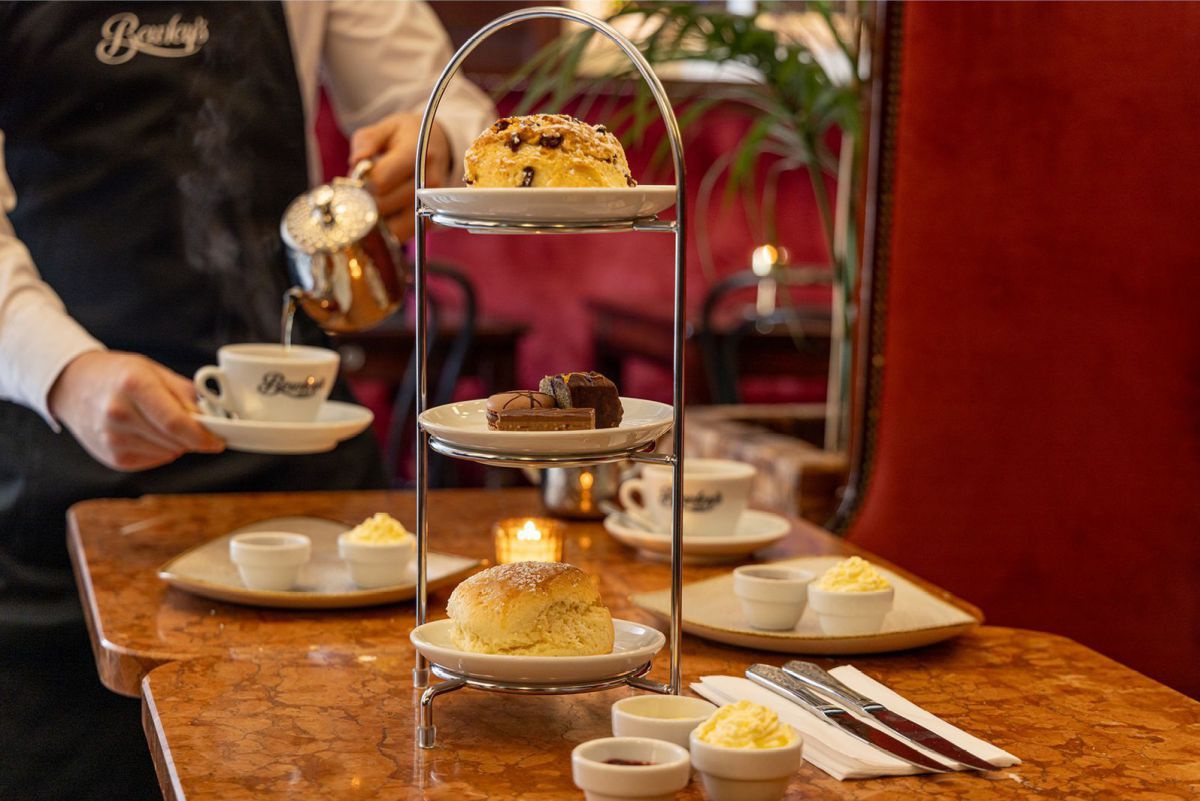Auf zwei nebeneinanderstehenden Kaffetischchen sind Scones, Creme und Marmelade sowie andere Mehlspeisen angerichtet. Ein Kellner schenkt gerade heißes Wasser aus einem silbernen Kännchen in eine Teetasse.