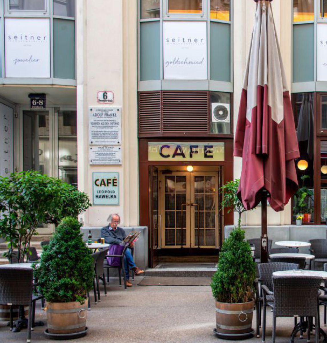 Wiener Kaffehaus von außen, ein Gast sitzt zeitungslesend im Schanigarten. Über dem Eingangstor steht groß 