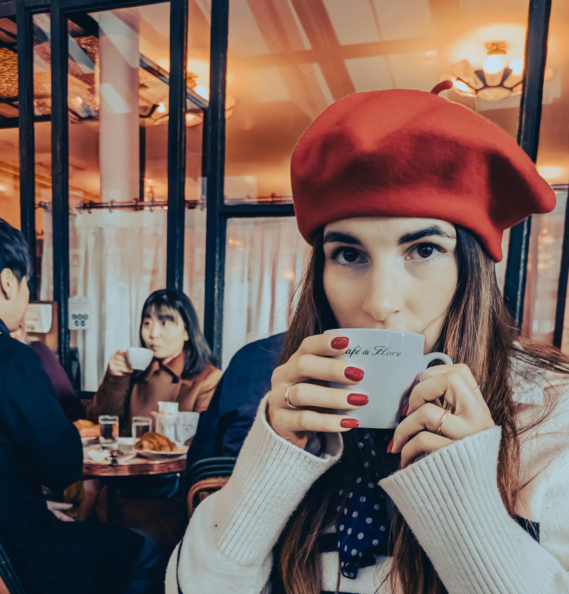 Im inneren eines Cafès: Frau mit langen dunklen Haaren, dunklen Augen und rotem Béret auf dem Kopf, trinkt aus einer weißen Tasse mit der Aufschrift 