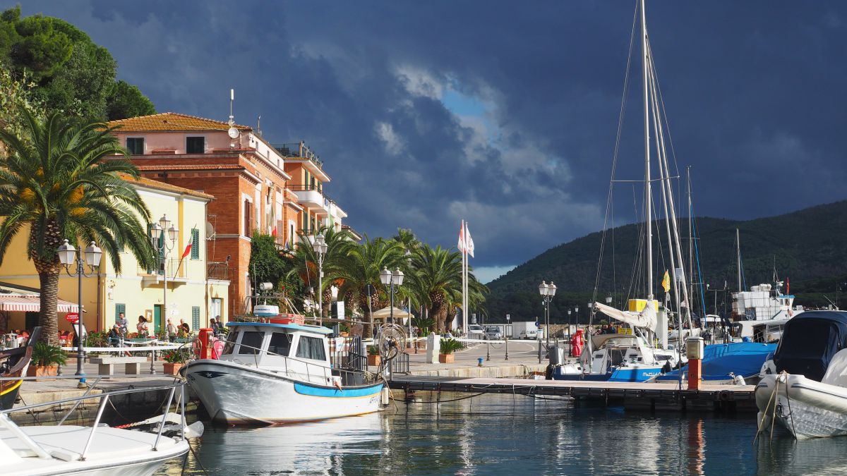 Sonnenbeschienener Hafen, Boote am Wasser, Prachtvolle Gebäude, Palmen, dunkle Wolken ziehen am Himmel auf.