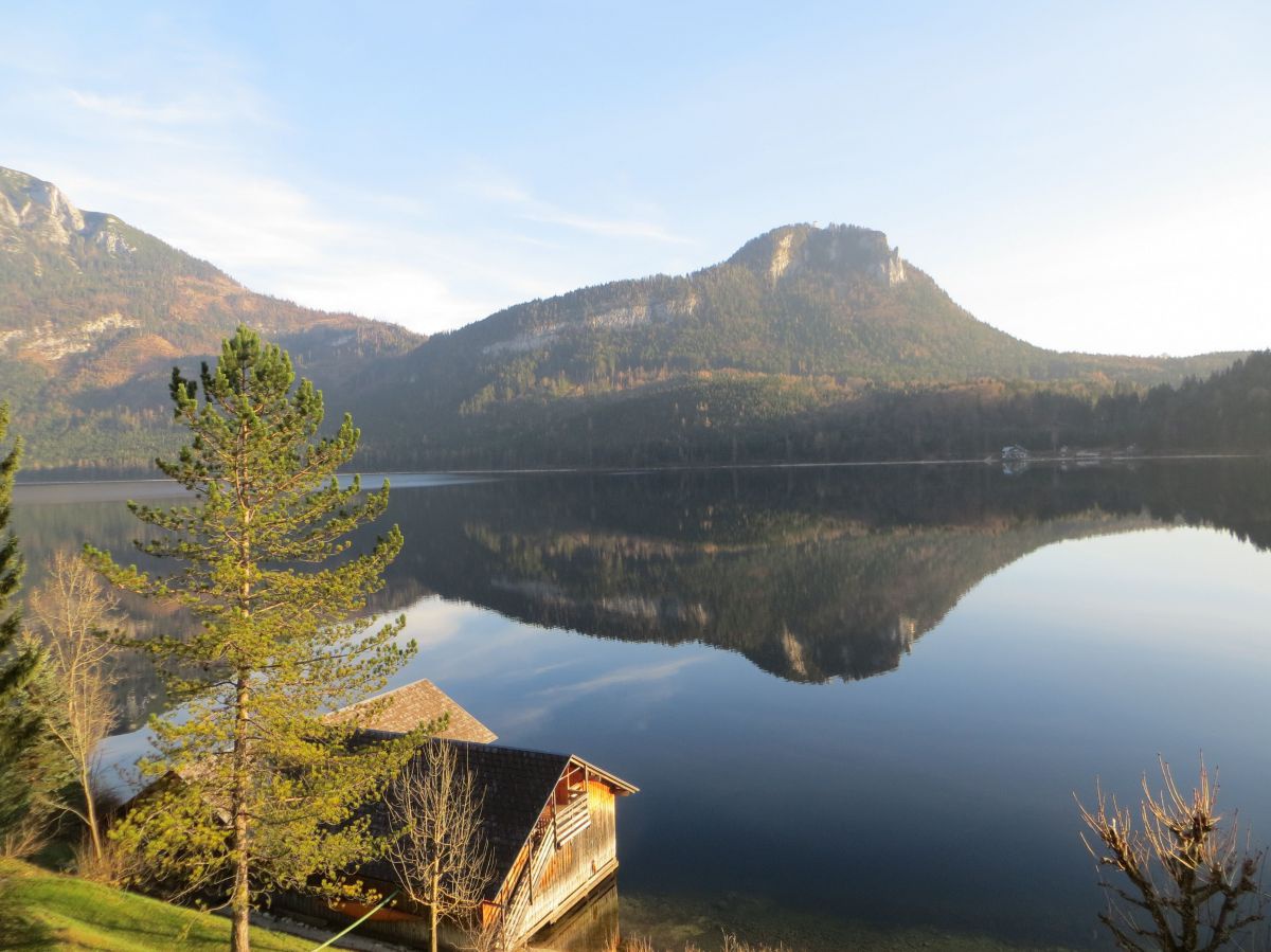 Spiegelglatte Wasserfläche zwischen bewaldeter Berglandschaft. Links vorne im Bild ein hölzernes Bootshaus unter hohen Nadelbäumen.