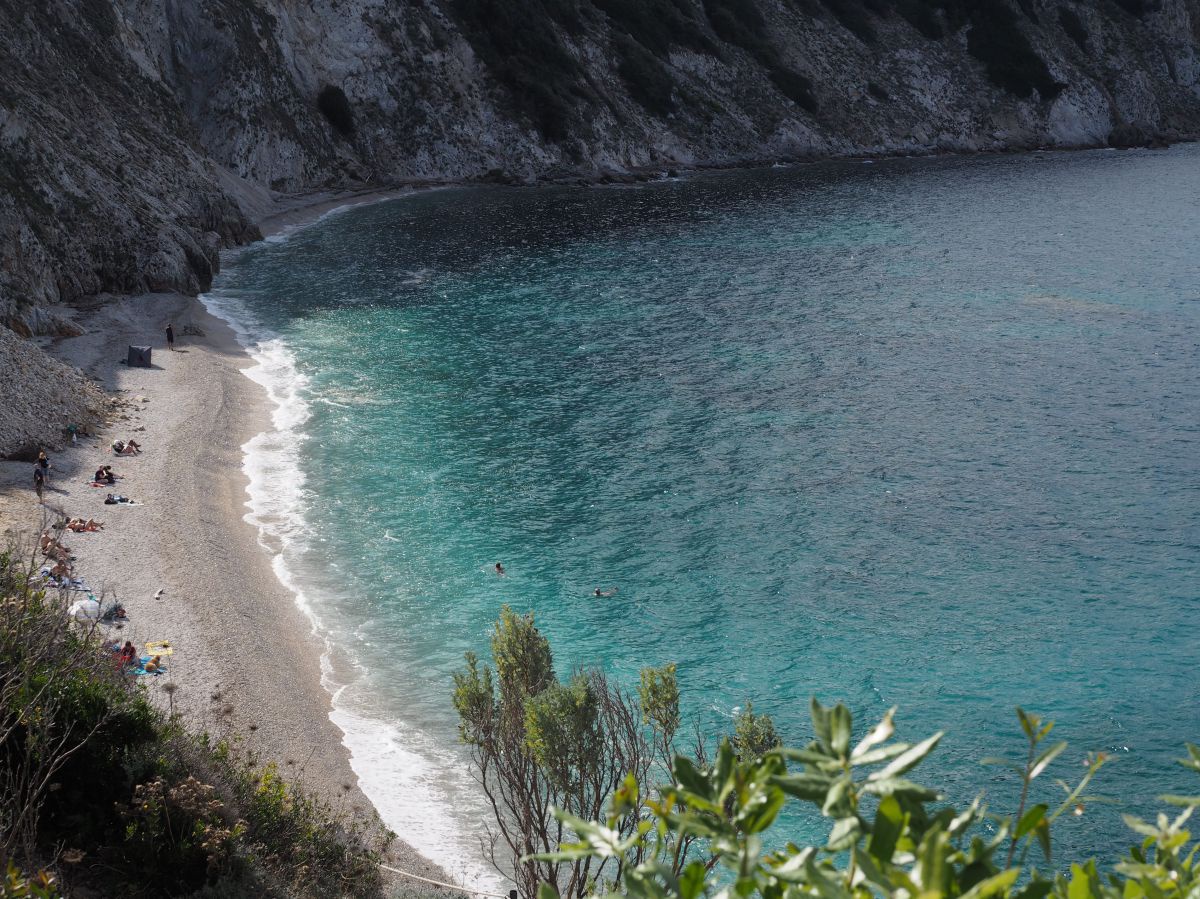 Blick von oben in eine Felsige Bucht mit kleinem Sandstrand. Das Wasser in der Bandung schimmert türkisblau, dazu die weiße Gischt.