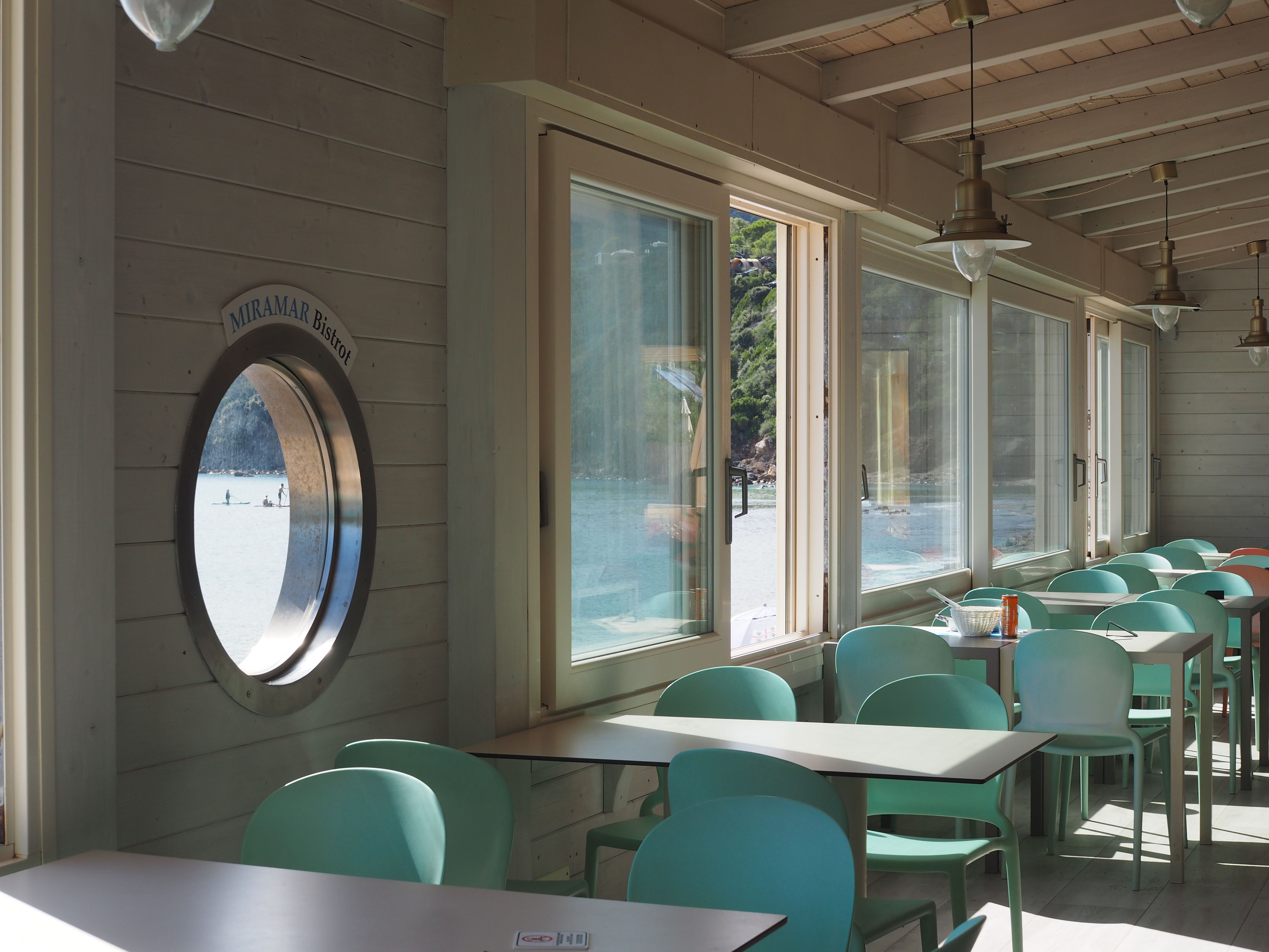 Innenbereich des Bistrot: weiße Tische mit türkisblauen Kunststoffstühlen, große Fenster lassen viel Licht herein, ein rundes Fenster erinnert an ein Bullauge.
