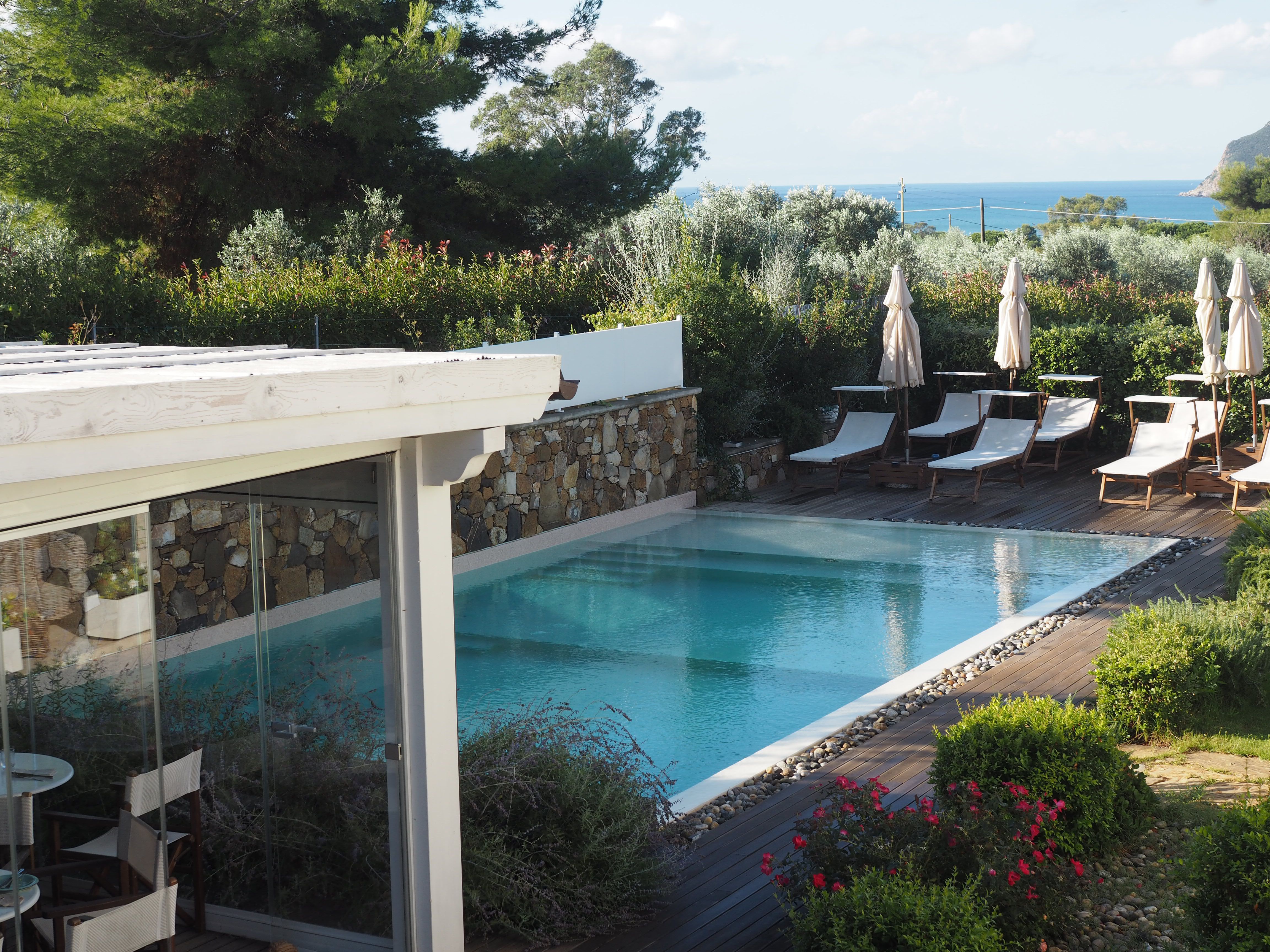 Blick auf einen Garten mit Pool, weißen Liegen und Sonnenschirmen. rundherum Bäume und blühende Sträucher.