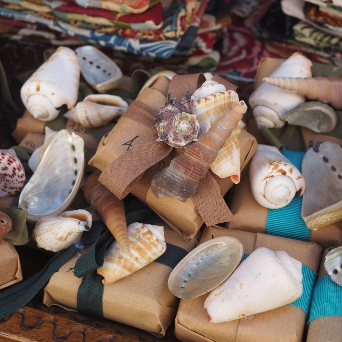 Kleine in Packpapier eingepackte Mitbringsel, mit Muscheln dekoriert, dahinter bunte Stoffe. 