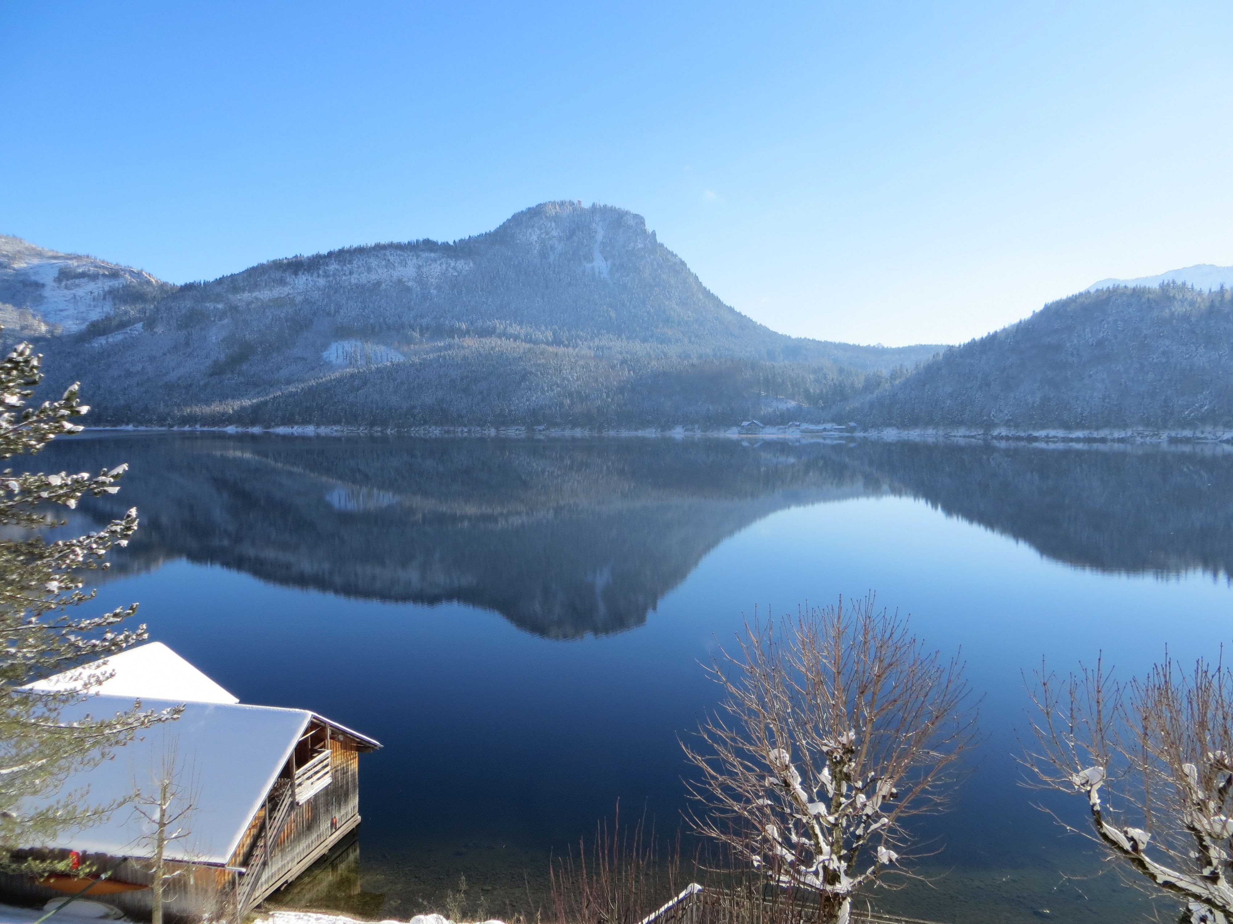 Spiegelglatte Wasserfläche zwischen bewaldeter Berglandschaft. Links vorne im Bild ein hölzernes Bootshaus unter hohen Nadelbäumen. Auf den Bergen, den Bäumen und den Dächern liegt Neuschnee.