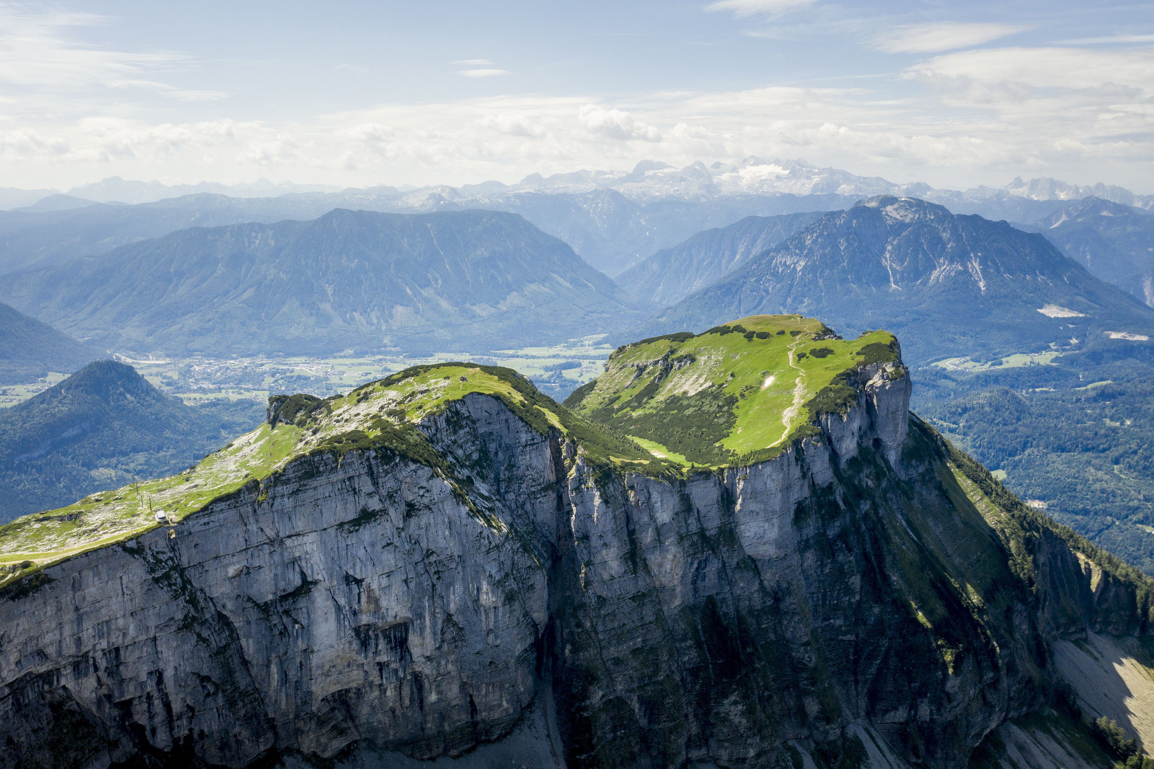 Drohnenaufnahme eines Berges. Das Plateau ist von hell- und dunkelgrüner Vegetation bedeckt, seitlich fällt die schroffe Felswand fast senkrecht hunderte Meter ins Tal. Im Hintergrund sieht man durch leicht trübe, diesige Luft weiteres Gebirge.