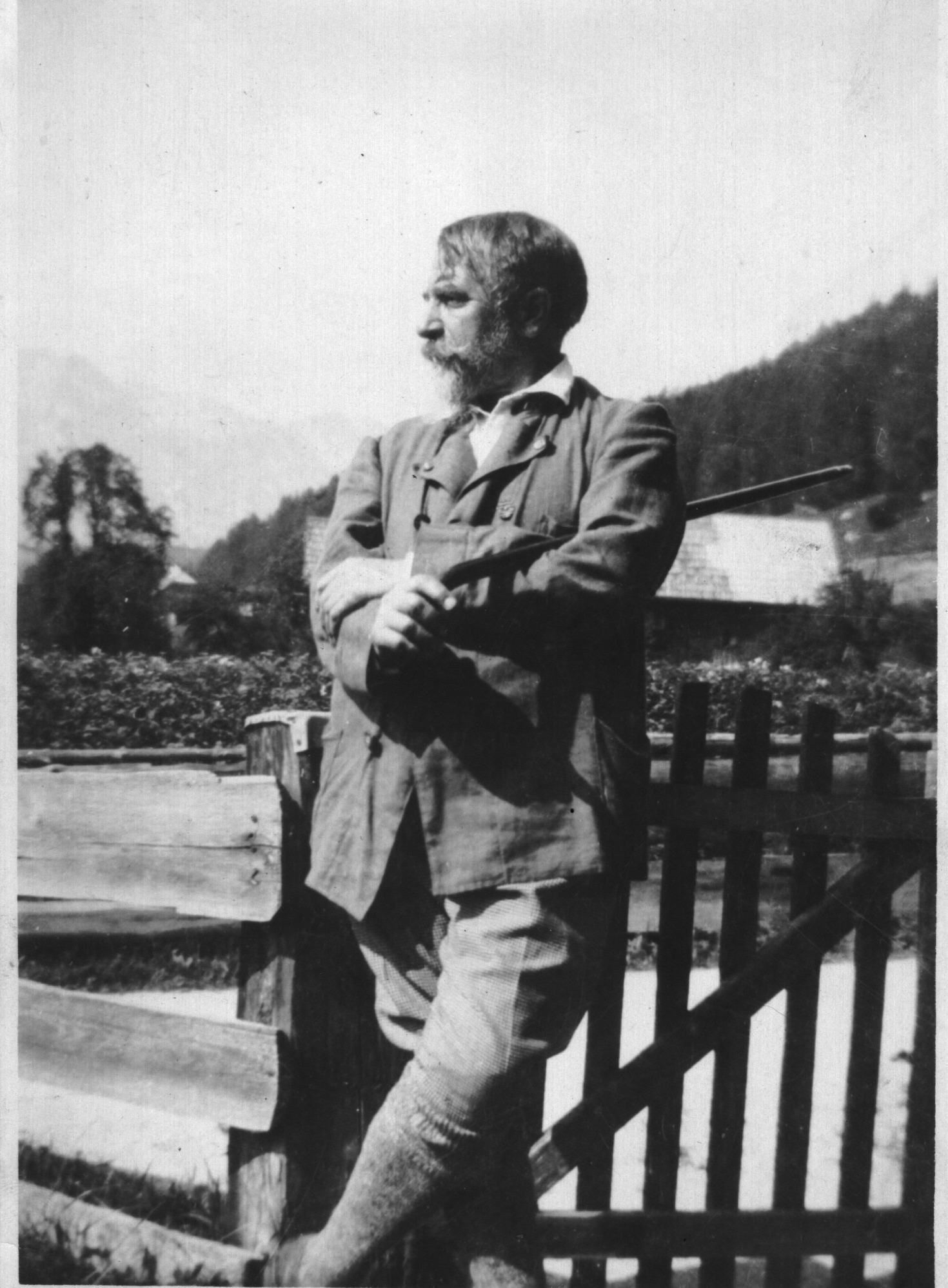 Schwarz-Weiß-Foto zeigt Mann mit buschigem Bart, in Wanderkleidung an einem Zaun lehnend, den Gehstock lässig unter die Achsel geklemmt und in die Ferne blickend.