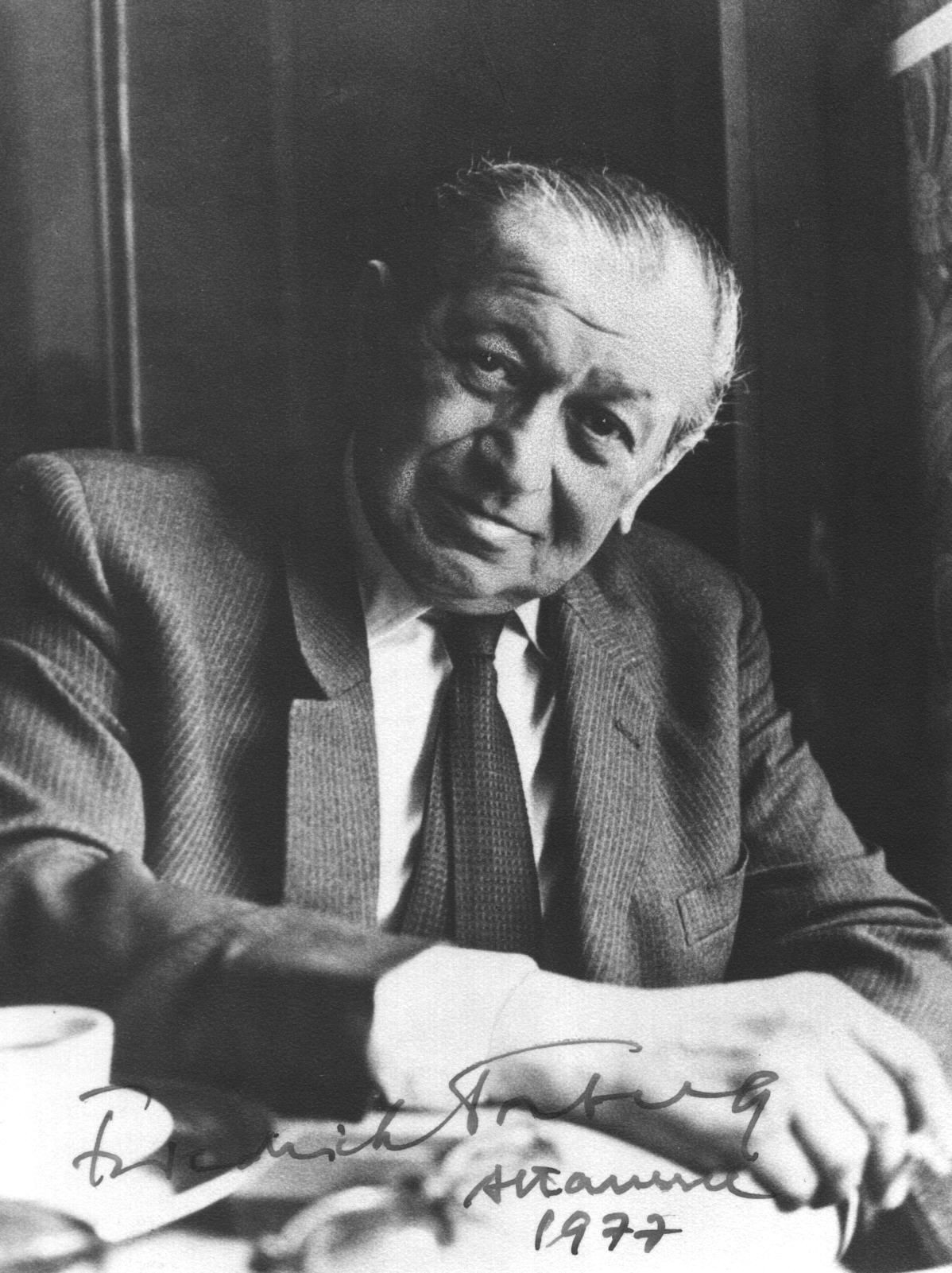 Schwarz-Weiß-Foto zeigt älteren Herrn mit zurückgekämmtem, schon etwas schütterem Haar am Kaffehaustisch sitzend und freundlich in die Kamera blickend. Am unteren Rand des Fotos sieht man die Signatur des Schriftstellers mit Ort und Jahreszahl.