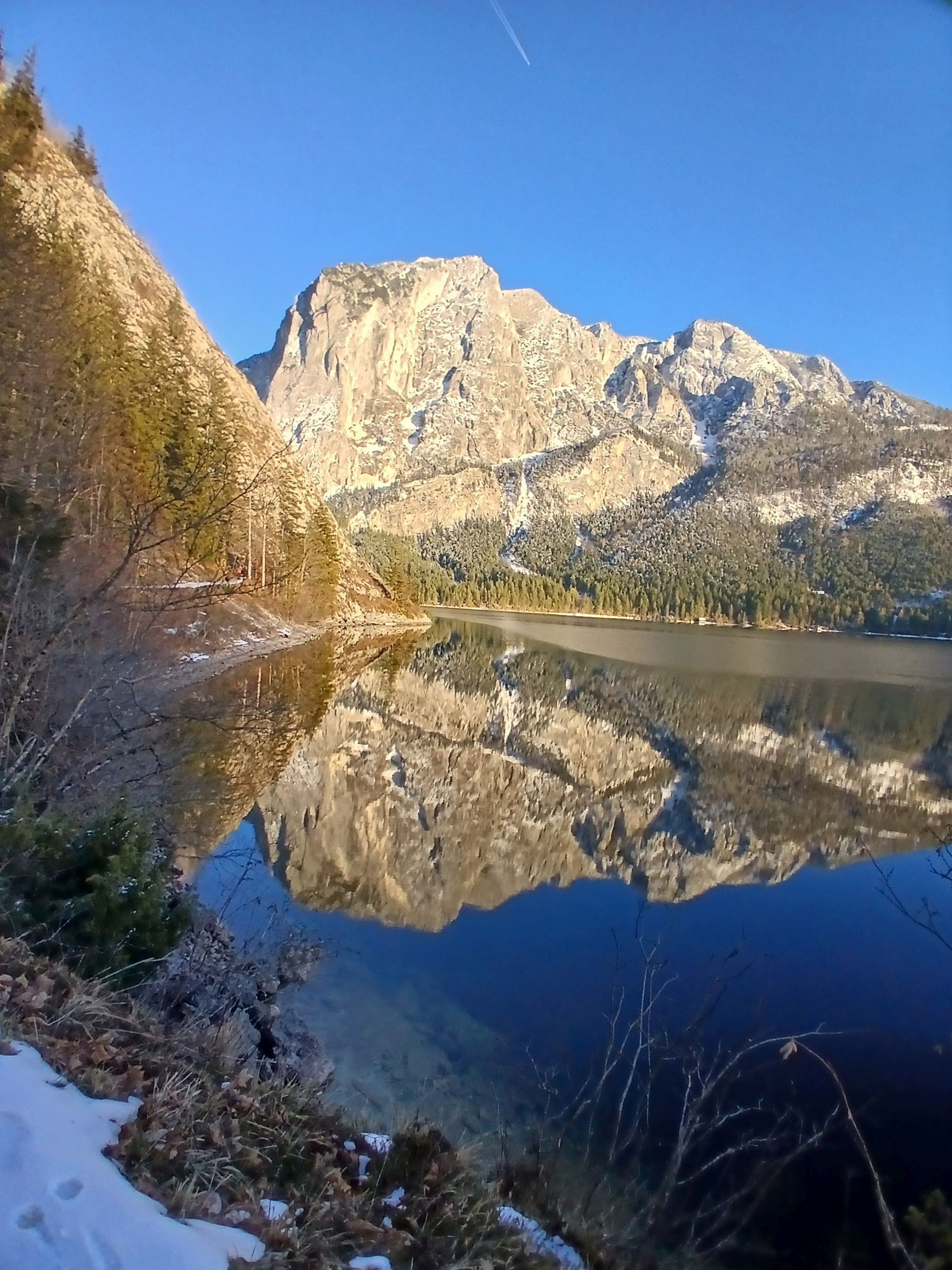 Es ist ein sonniger Tag, in der glatten Wasserfläche des Sees spiegelt sich das riesige Bergmassiv. Am Seeufer liegt ein wenig Schnee, in dem kleine Tierspuren zu sehen sind.
