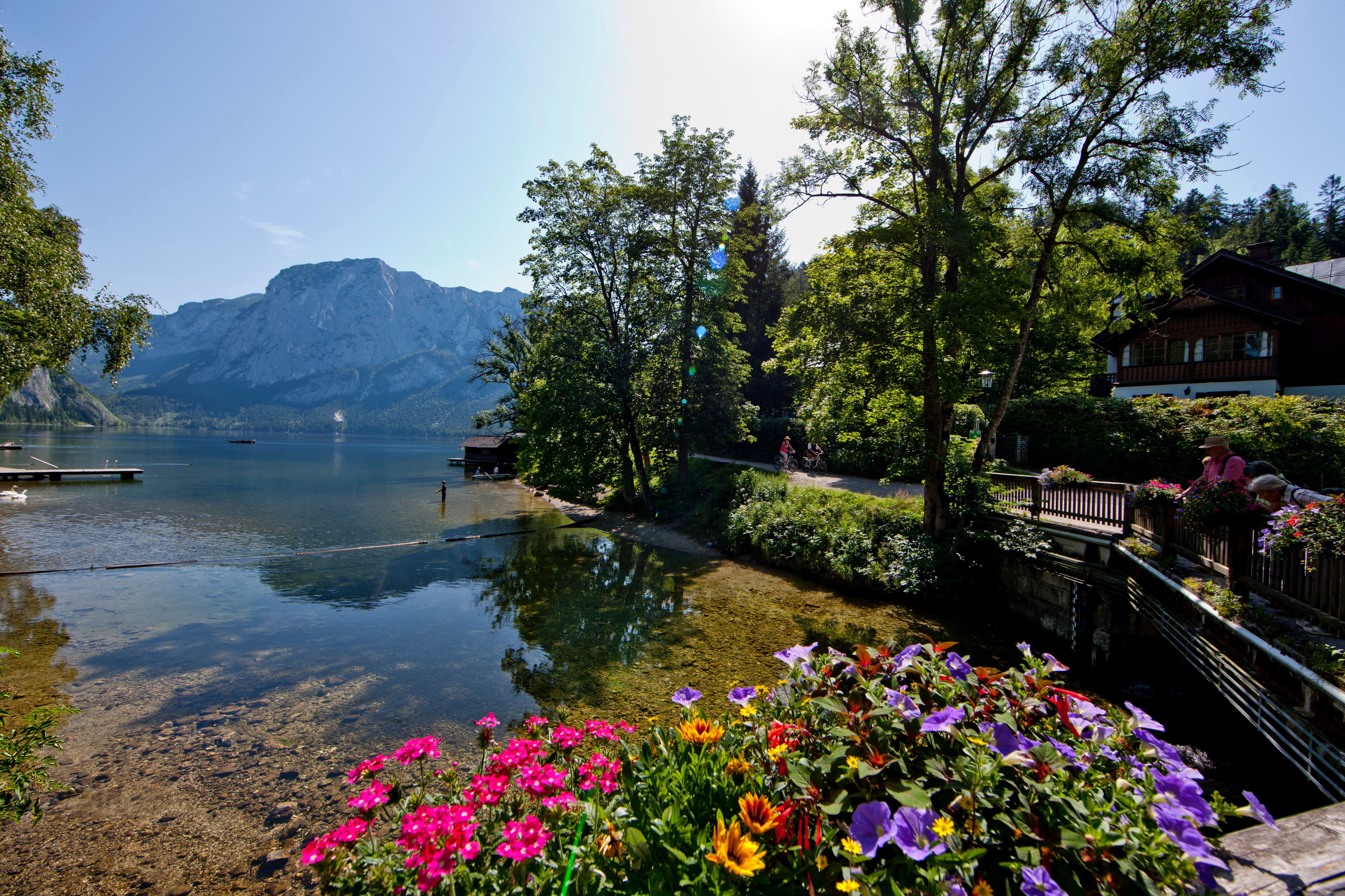 Die Straße am Seeufer führt hier über eine Brücke und ist mit einem Holzgeländer gesäumt, auf dem in Blumenkisten viele bunte Balkonblumen blühen. Ein paar Sommergäste stehen am Geländer und blicken auf den See.