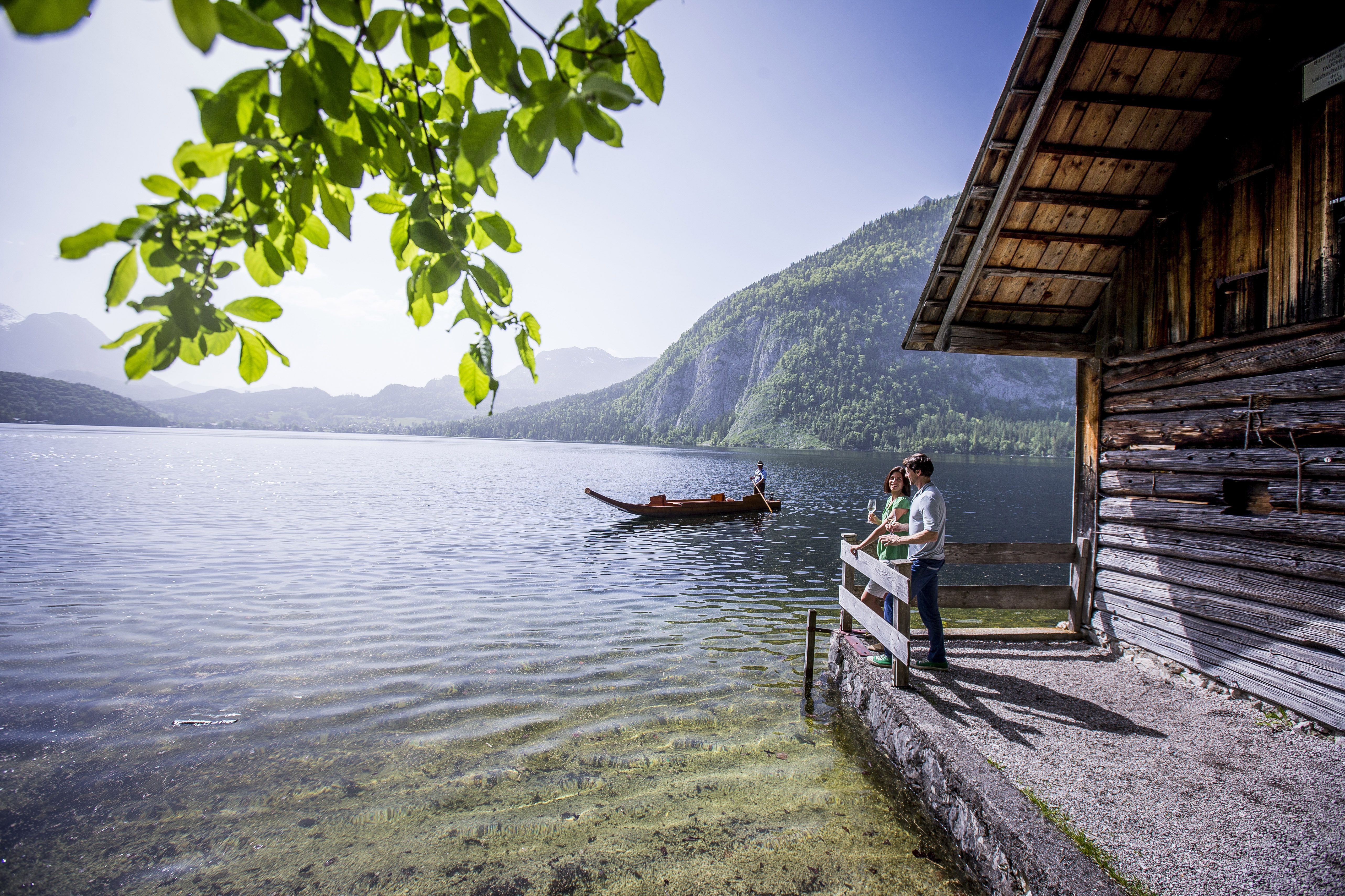 Vor einem Bootshaus steht ein Paar am Geländer und blickt auf den See, dessen Wasserfläche leicht gewellt ist. Jemand fährt mit einer Zille über den See. Im Hintergrund Gebirge, im Vordergrund hängt von oben ein leuchtend grün belaubter Zweig eines Baumes
