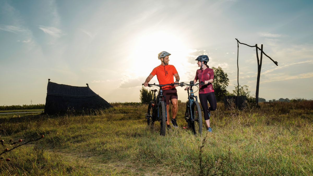 2 Radfahrer bei Sonnenuntergang im Gras stehend, dahinter ein typischer Schöpfbrunnen.