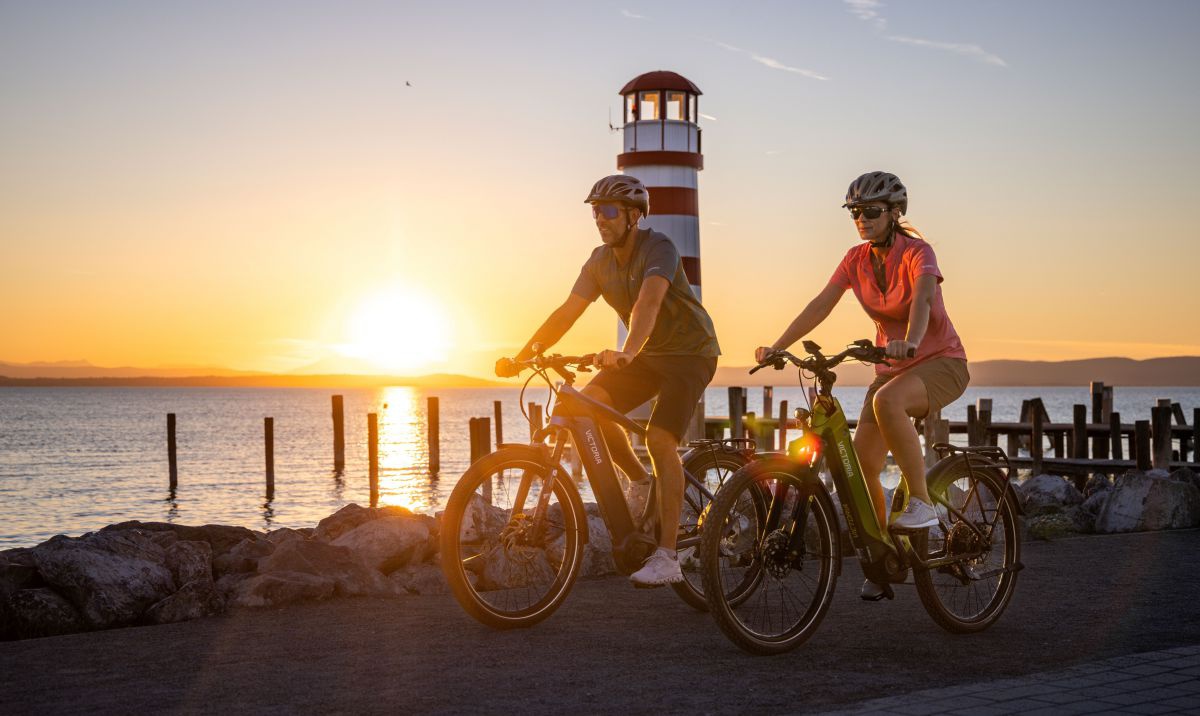 Mann und Frau Radeln am See, vorbei an einem Leuchtturm, im Hintergrund geht die Sonne unter.