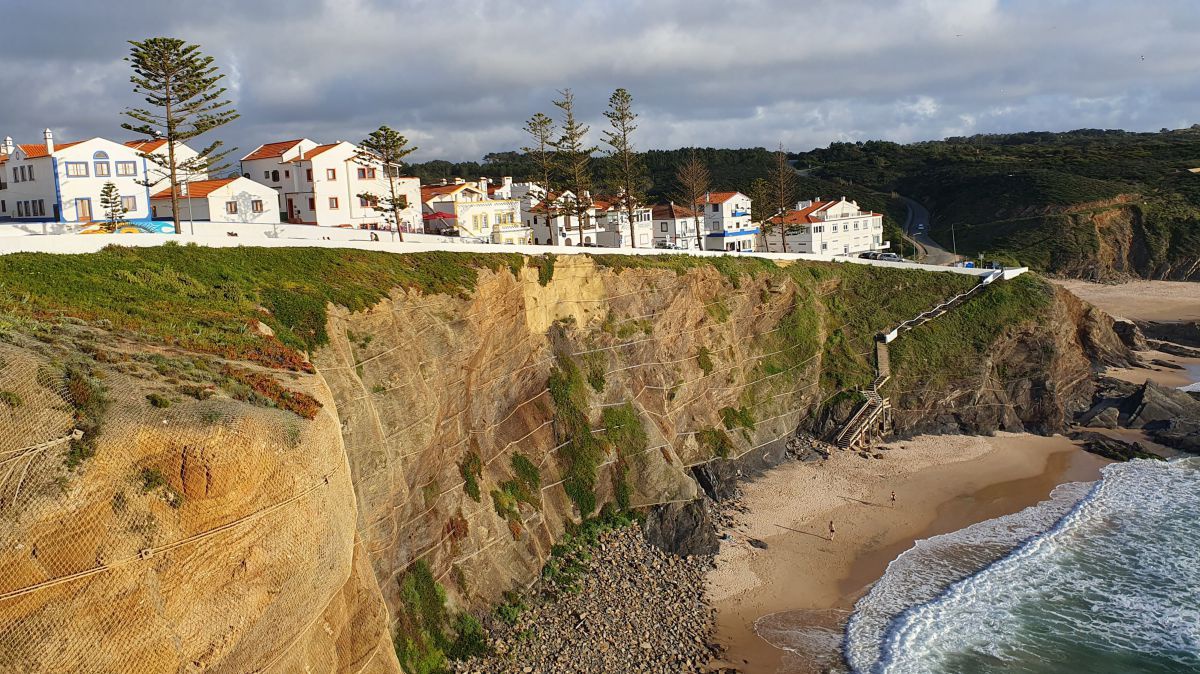Weiß glänzende Häuser auf einem Felsen, darunter eine Bucht und Meereswellen.