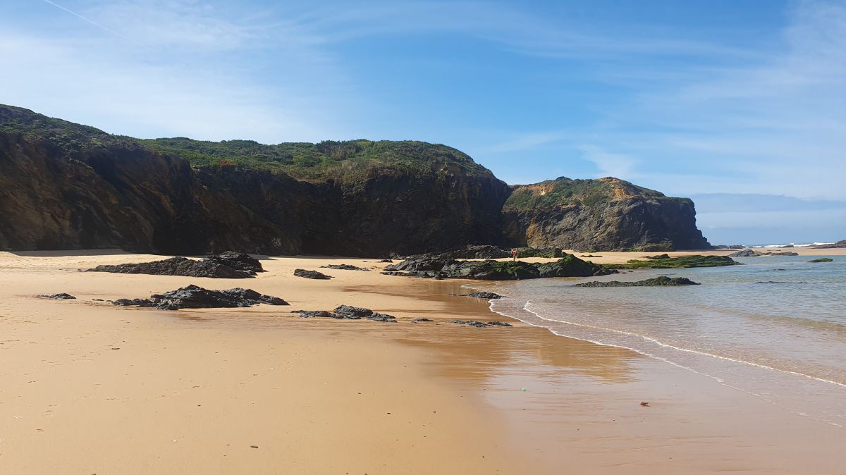 Sandstrand, dahinter Felsen, das Meer ist ruhig.