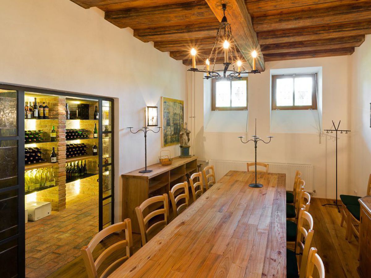 Rustikaler Raum mit einem länglichen Tisch, links eine beleuchtete Vitrine mit Weinflaschen.