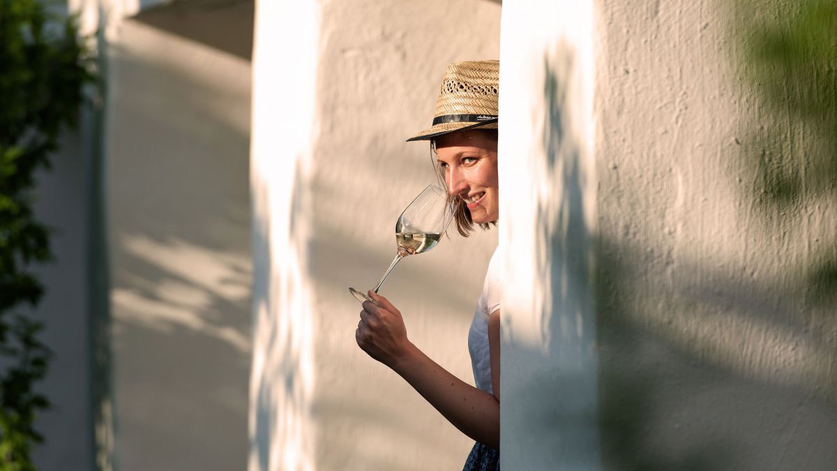 Frau mit Strohhut und Weinglas lehnt an einer Mauer.
