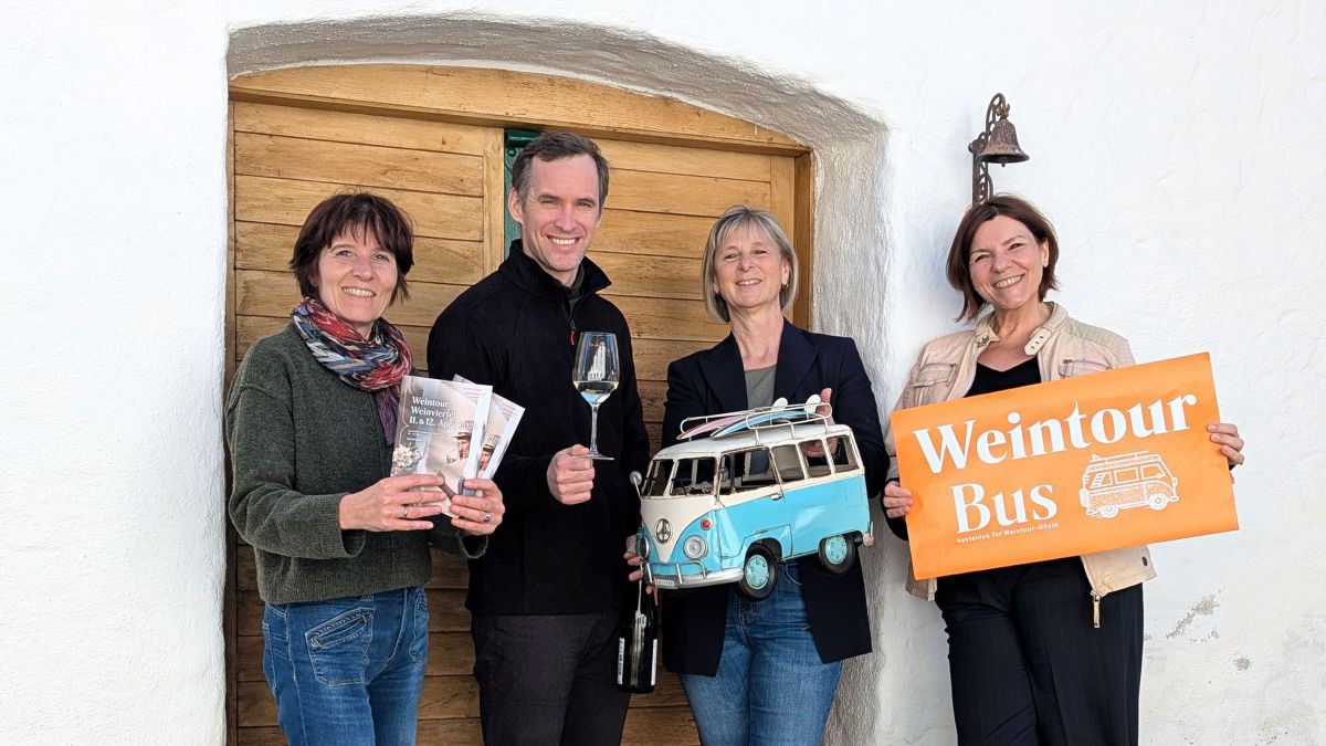 4 Personen vor der Tür in einen Weinkeller. Sie halten Prospekte, ein Weinglas, ein Modell eines VW-Busses und ein Schild 