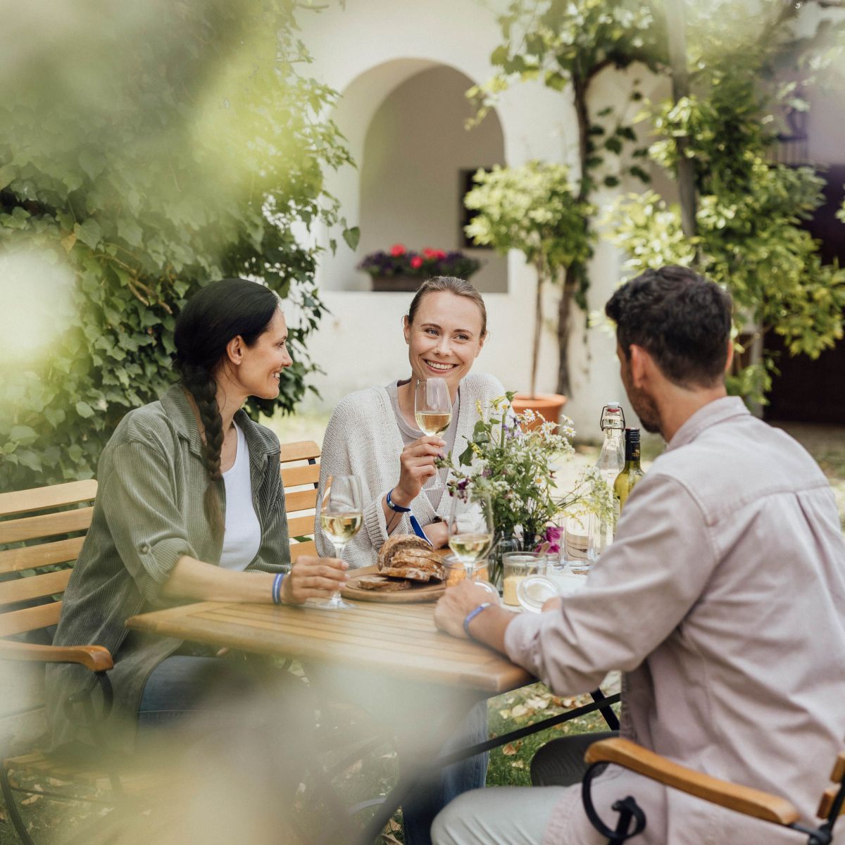 2 Frauen und ein Mann sitzen im Garten eines Heurigenbetriebes bei einer Jause mit Wein.